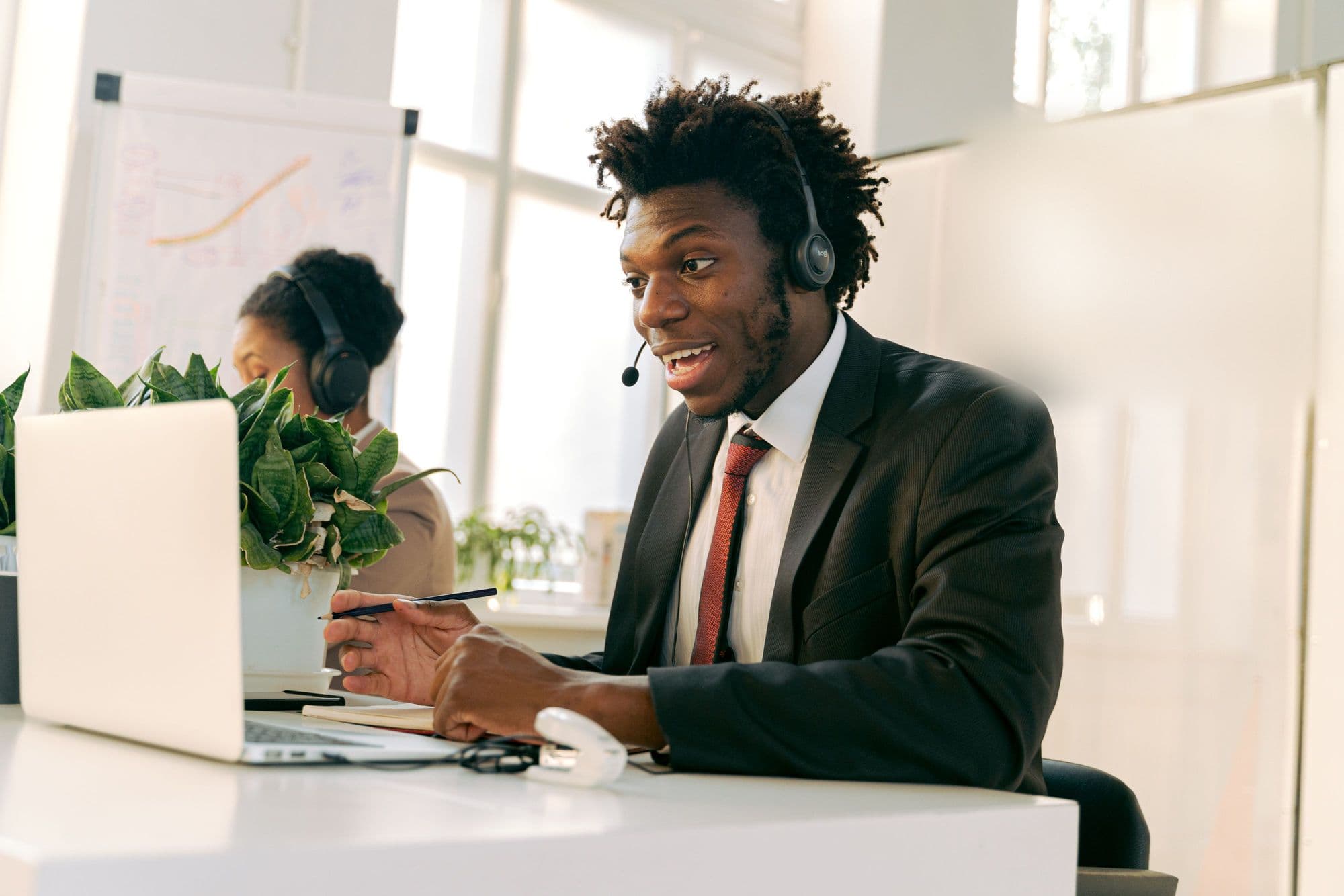 A smiling professional in a suit taking a call on a headset at his laptop